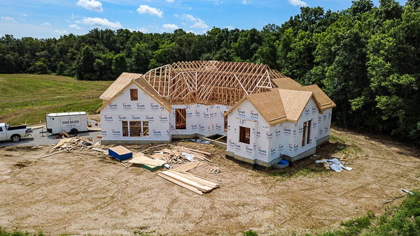 An aerial, eye-level shot captures a large house under construction in a rural, wooded area under a bright, partly cloudy sky. The house, positioned in the center, features a complex wooden roof frame with exposed trusses, and its exterior walls are wrapped in white Tyvek weather-resistant barriers. Portions of the roof are covered with plywood sheathing, while other parts show the skeletal structure of the wooden rafters. The building sits on a concrete foundation in a clearing of dry, light-brown dirt marked with tire tracks. Scattered around the site are construction materials, including neat piles of wooden planks, loose plywood sheets, and scraps of lumber. To the left of the house, a white pickup truck is parked next to a white enclosed utility trailer labeled "RENEW BUILDERS." A dense wall of lush green trees surrounds the construction clearing on the right and in the background, while a grassy field extends to the left.