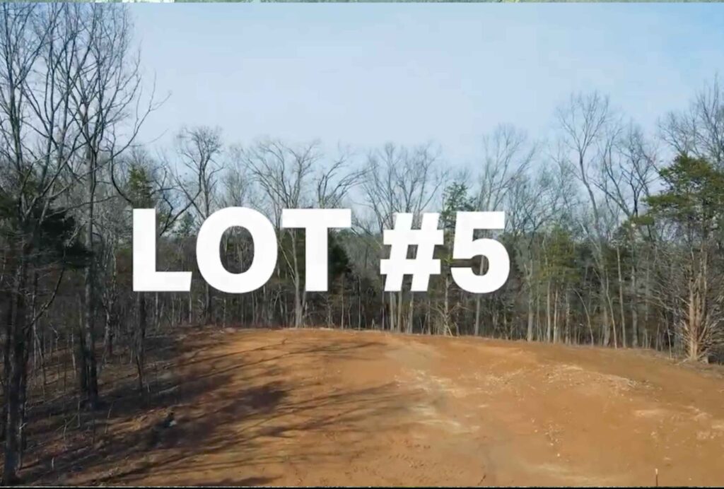 Alt text: An elevated view of a cleared, dirt real estate lot numbered "LOT #5" in large white text. The foreground shows a graded patch of brown earth, bordered by a dense line of bare winter trees under a clear, light blue sky.