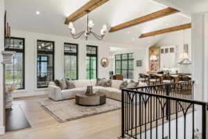 A bright, open-concept living room featuring a white sectional sofa, a round wooden coffee table, and exposed wood ceiling beams. The space seamlessly flows into a kitchen with an island and a dining area visible through large black-framed windows.