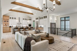 "An open-concept great room combining a living area and kitchen under a high, vaulted ceiling with exposed wooden beams. In the foreground, a cream-colored sectional sofa is arranged with patterned accent pillows and a dark throw blanket, facing a round wooden coffee table on a textured area rug. A large black wrought-iron chandelier hangs from the ceiling. In the background, the kitchen features white upper cabinets, light wood lower cabinetry, a large center island with barstools, and a tall wood-framed curio cabinet with wire mesh detailing on the far left. A black metal stair railing sits to the right."