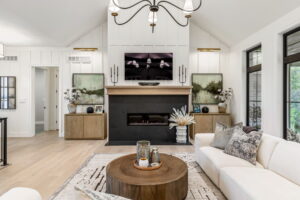 A modern living room centering on a large fireplace with a black surround and light wood mantel. A TV is mounted above the mantel, flanked by two identical abstract paintings resting on low wood cabinets under warm accent lighting.
