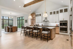 A spacious kitchen showcasing a light wood island with four modern barstools, white upper cabinetry, and integrated appliances. A cozy dining table with leather chairs sits adjacent to the kitchen, framed by large windows looking out to a wooded view.