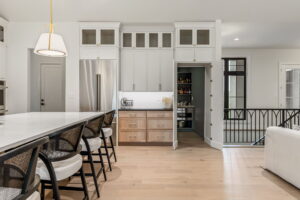 "A bright kitchen area showcasing a hidden pantry installation. The main wall features white upper cabinets, glass-and-wire-mesh accent cabinets at the top, and a stainless steel refrigerator next to a light wood perimeter base with drawers. One of the large white cabinet doors is swung wide open, revealing a secret, walk-in pantry room equipped with dark gray shelving, small appliances, and bottles inside. In the foreground, a white kitchen island counter is lined with black wooden barstools featuring woven cane backs and white cushions."