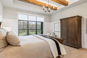 A spacious master bedroom with a large bed dressed in beige linens. The ceiling features a tray design with three thick, natural wood beams and a central black iron chandelier. In the background, a large black-trimmed triple window with transom panels overlooks a grassy backyard and trees. A tall, rustic dark wood armoire stands against the right wall, which is finished with white shaker wainscoting paneling.
