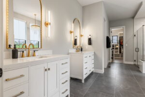 A modern master bathroom featuring dark grey slate floor tiles and two separate white vanities. Each vanity has a white quartz countertop, a modern gold faucet, and a large arch-shaped mirror with a gold frame. Vertical gold and glass sconces illuminate the space. The room opens directly into a brightly lit walk-in closet with built-in shelving visible in the background."