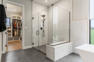 "A luxury walk-in shower with a clear glass enclosure and a dark grey mosaic tile floor. The walls are lined with large-format white marble tiles with subtle grey veining. Inside, there is a black rainfall showerhead with a handheld attachment, and a low marble-capped bench sits at the front. The open doorway next to the shower leads directly into a hardwood-floored walk-in closet filled with hanging clothes."