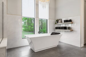 "A deep, modern white freestanding soaking tub centered beneath two large vertical windows that look out onto a green lawn and trees. The walls are tiled halfway with large white marble panels accented by a thin gold trim line. A delicate crystal chandelier hangs directly overhead, and two floating wooden shelves on the right wall hold rolled black towels and decorative items."