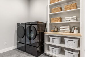 "A clean, functional laundry room featuring a dark charcoal gray matching washer and dryer set on the left. To the right, a large white built-in storage unit includes a light wood countertop for folding, open shelving holding woven wicker baskets and folded white towels, and lower compartments filled with white plastic laundry baskets. The floor is covered in large dark grey tiles."