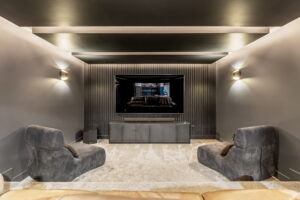 A cozy home theater room with dark walls, vertical wood slat accent paneling behind a large wall-mounted screen, subtle ambient ceiling lighting, and comfortable low-profile lounge chairs.