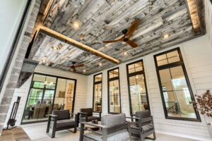 An alternative angle of the screened porch, highlighting the detailed weathered-wood plank ceiling with multiple recessed lights and two modern wood-blade ceiling fans. Large glass sliding doors and floor-to-ceiling windows show a glimpse of the indoor dining and living areas.