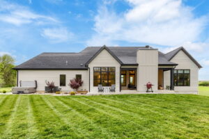 The back exterior of a modern ranch-style home featuring light siding, dark window frames, and a manicured green lawn. A patio area includes a pair of outdoor chairs, a hot tub on the left, and a red kettle grill near the screened porch entrance.