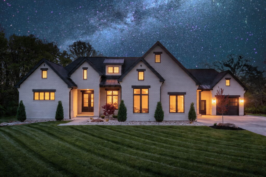 A dramatic nighttime shot of the home's front exterior, featuring a white brick facade with glowing warm light from the windows. The house sits under a vibrant, starry night sky with a clearly visible Milky Way galaxy.